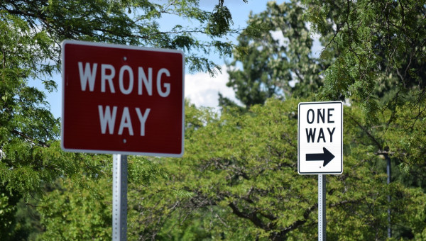 Fotografie zweier Straßenschilder nebeneinander. Auf dem vordergründigen Straßenschild heißt es 'Wrong Way'. Auf dem Straßenschild im Hintergrund heißt es 'One Way'