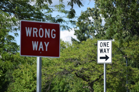 Fotografie zweier Straßenschilder nebeneinander. Auf dem vordergründigen Straßenschild heißt es 'Wrong Way'. Auf dem Straßenschild im Hintergrund heißt es 'One Way'
